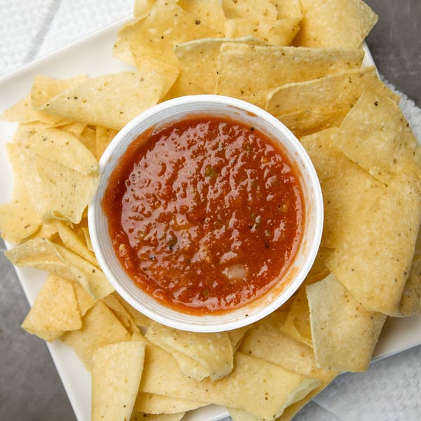 Overhead view of a white bowl of red salsa surrounded by golden tortilla chips on a white plate.